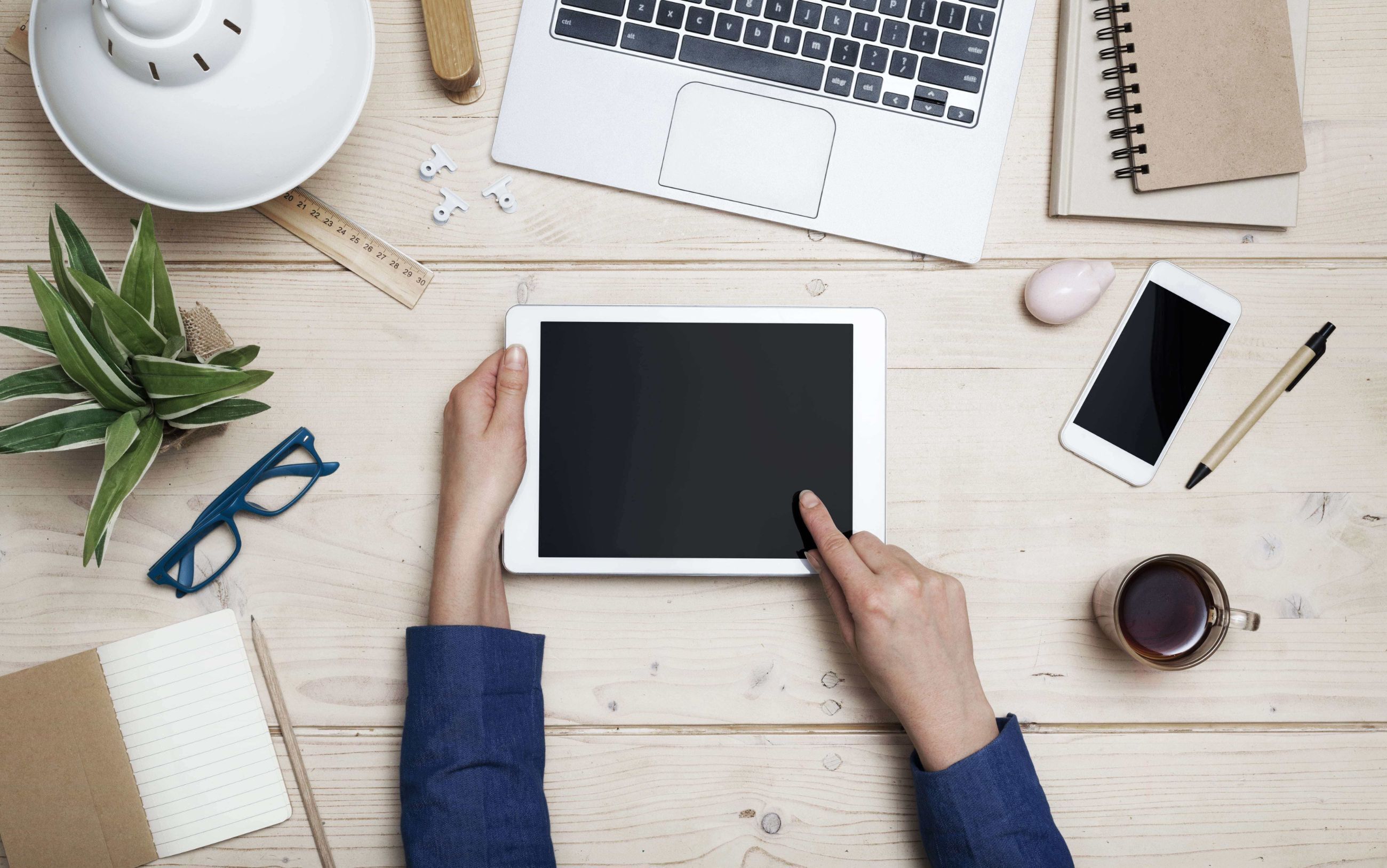 A pair of hands holding a tablet on a light wood desk surrounded by office equipement