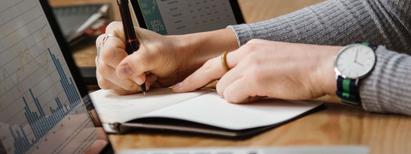 A photo of a person writing notes with two laptops on a table