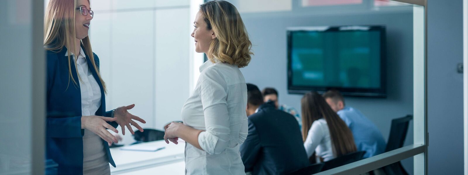 A photo through a glass wall of people talking in a meeting room with a television on the wall