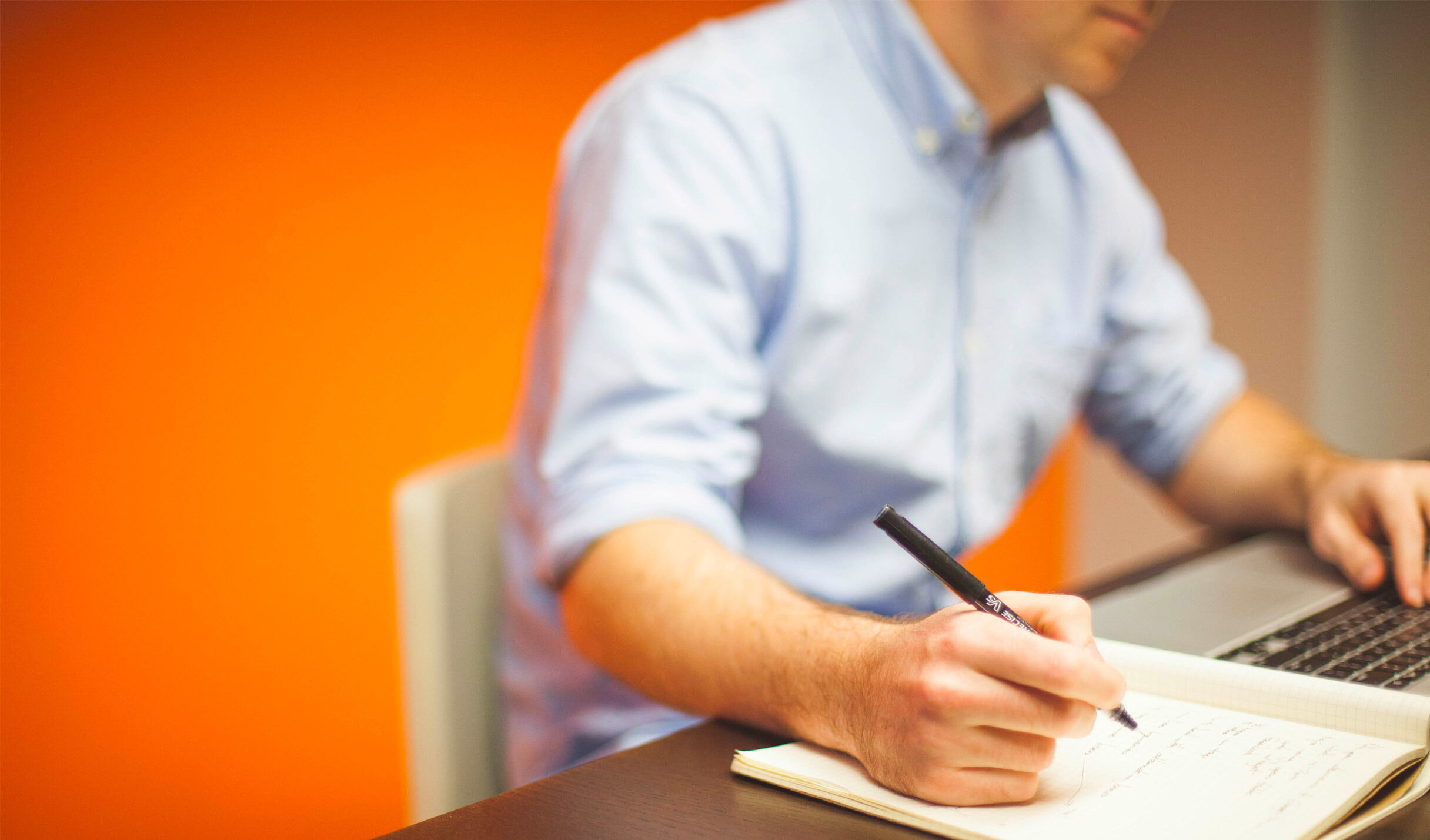 A person sitting at a desk writing notes and looking at a laptop