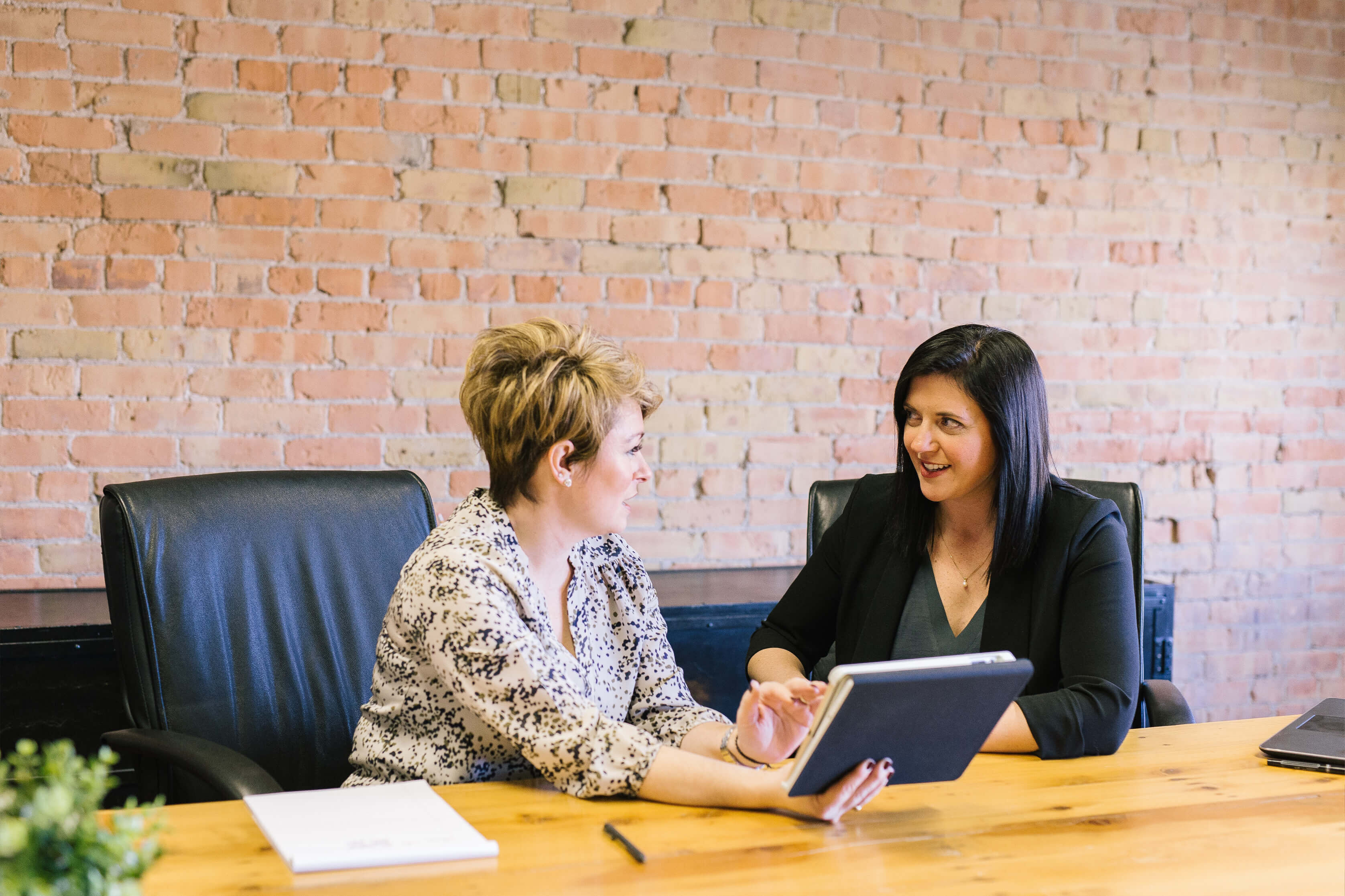 Two people talking sat around an oak desk in an office with bare brick walls