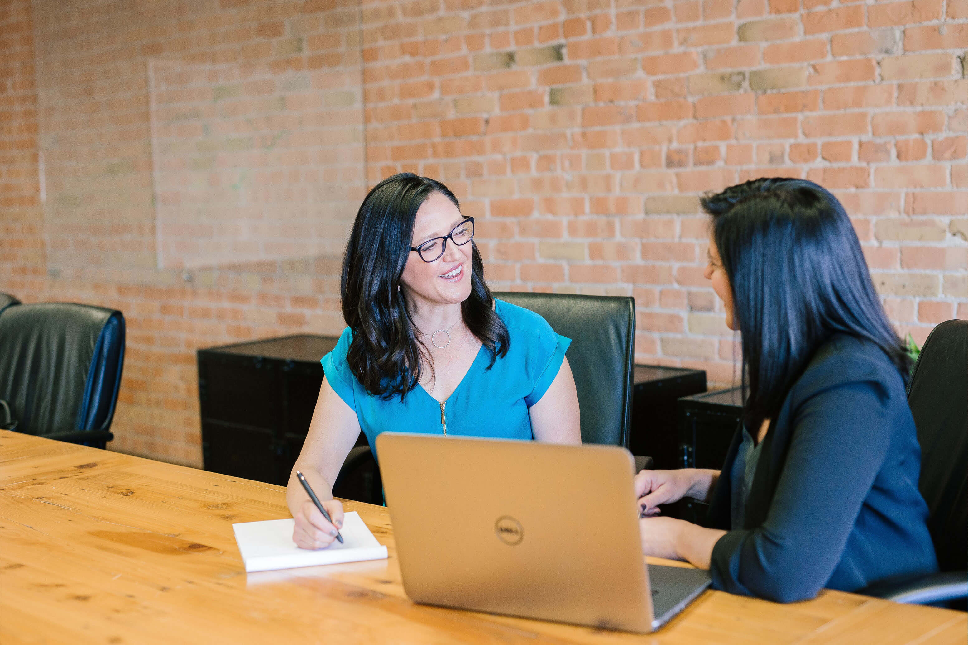 Two women talking in an office, light oak table, exposed brick wall, black chairs