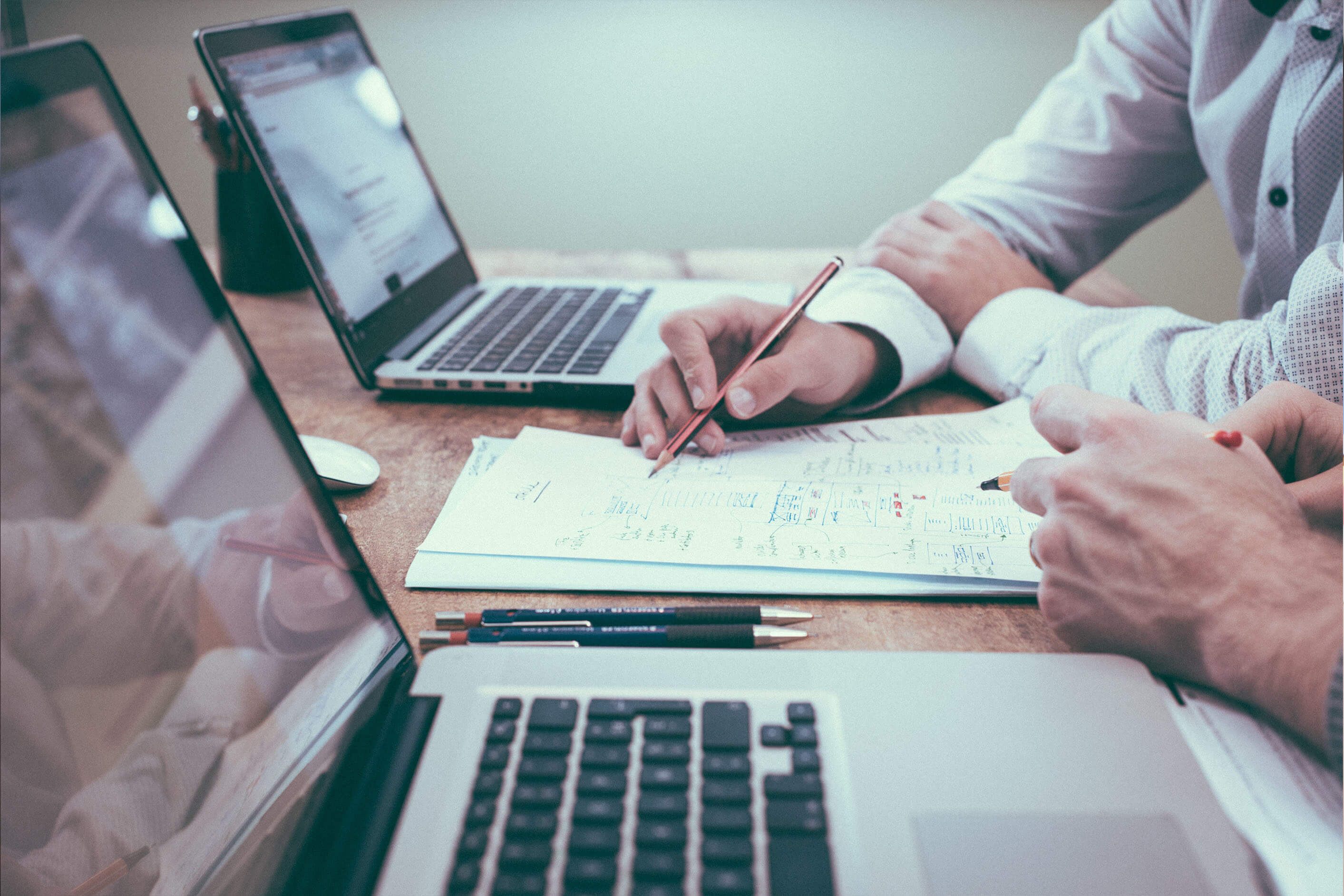 Photo of two laptops on a desk, two people looking at notes on paper