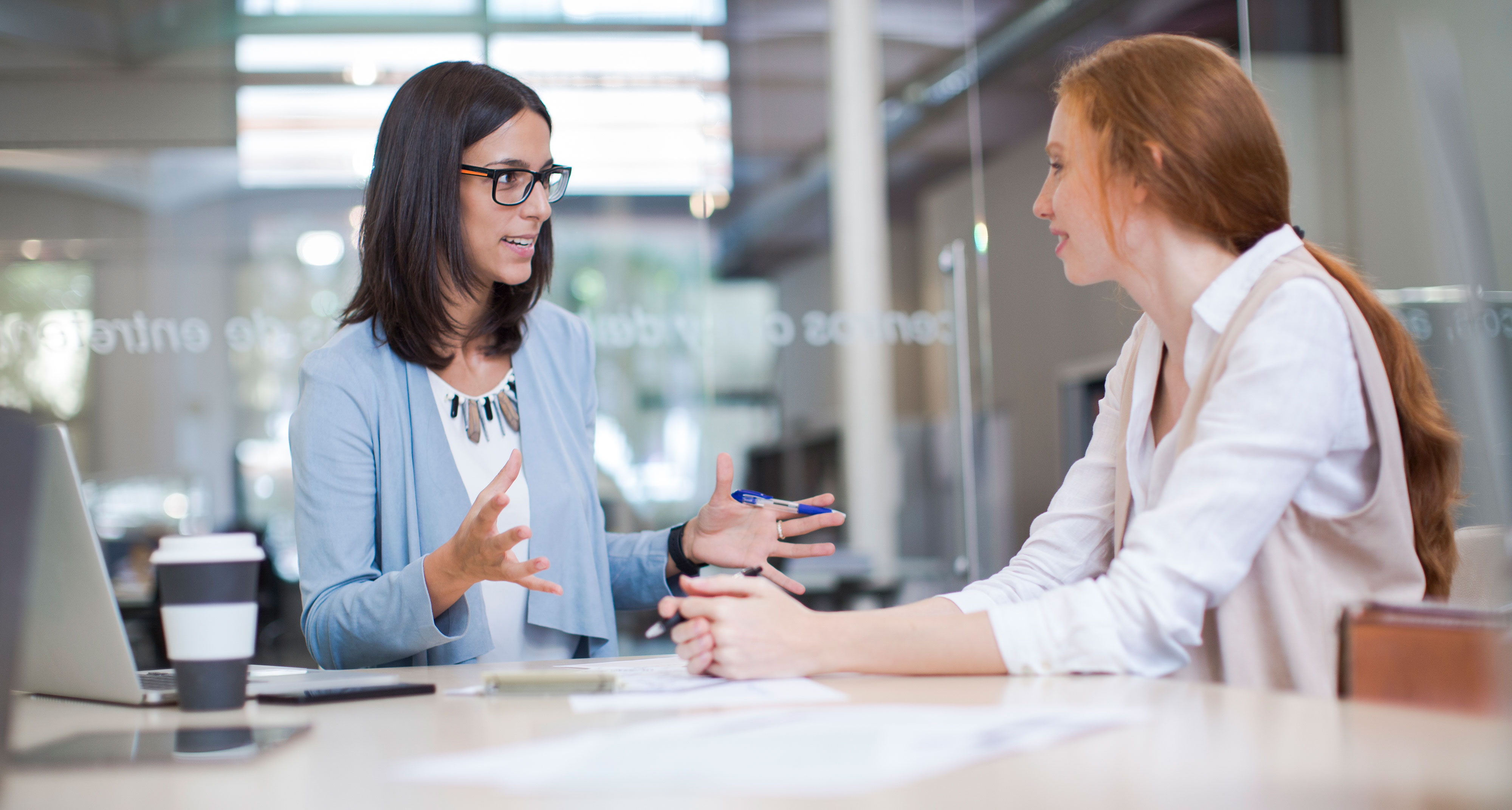 Two women talking in an office meeting room glass walls and door, white table, bright lighting